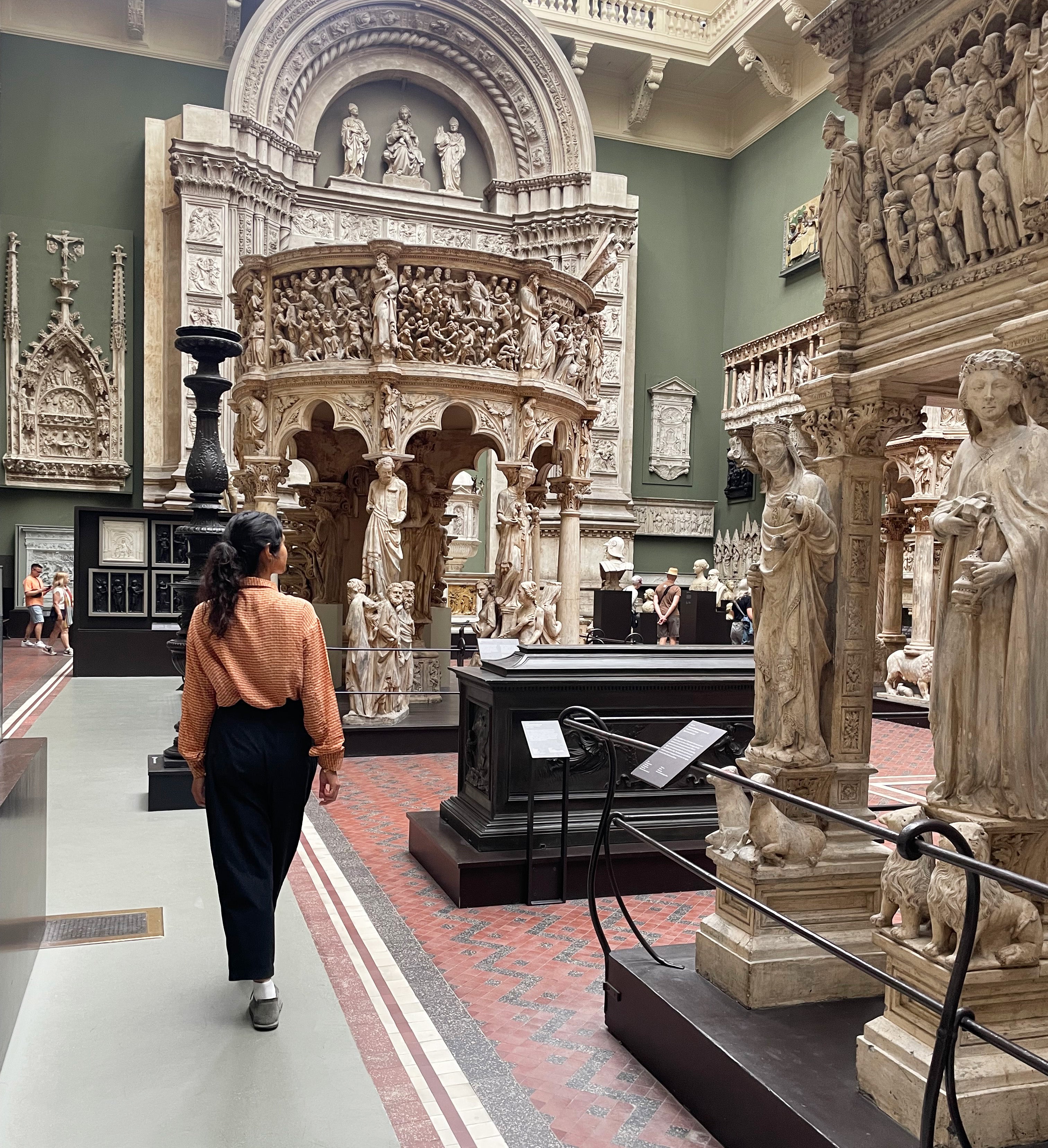 A woman in a rust-orange shirt and dark trousers walks through a large, ornate museum gallery filled with carved stone monuments and sculptures. It is one of the sculpture halls at the Victoria and Albert museum in London.