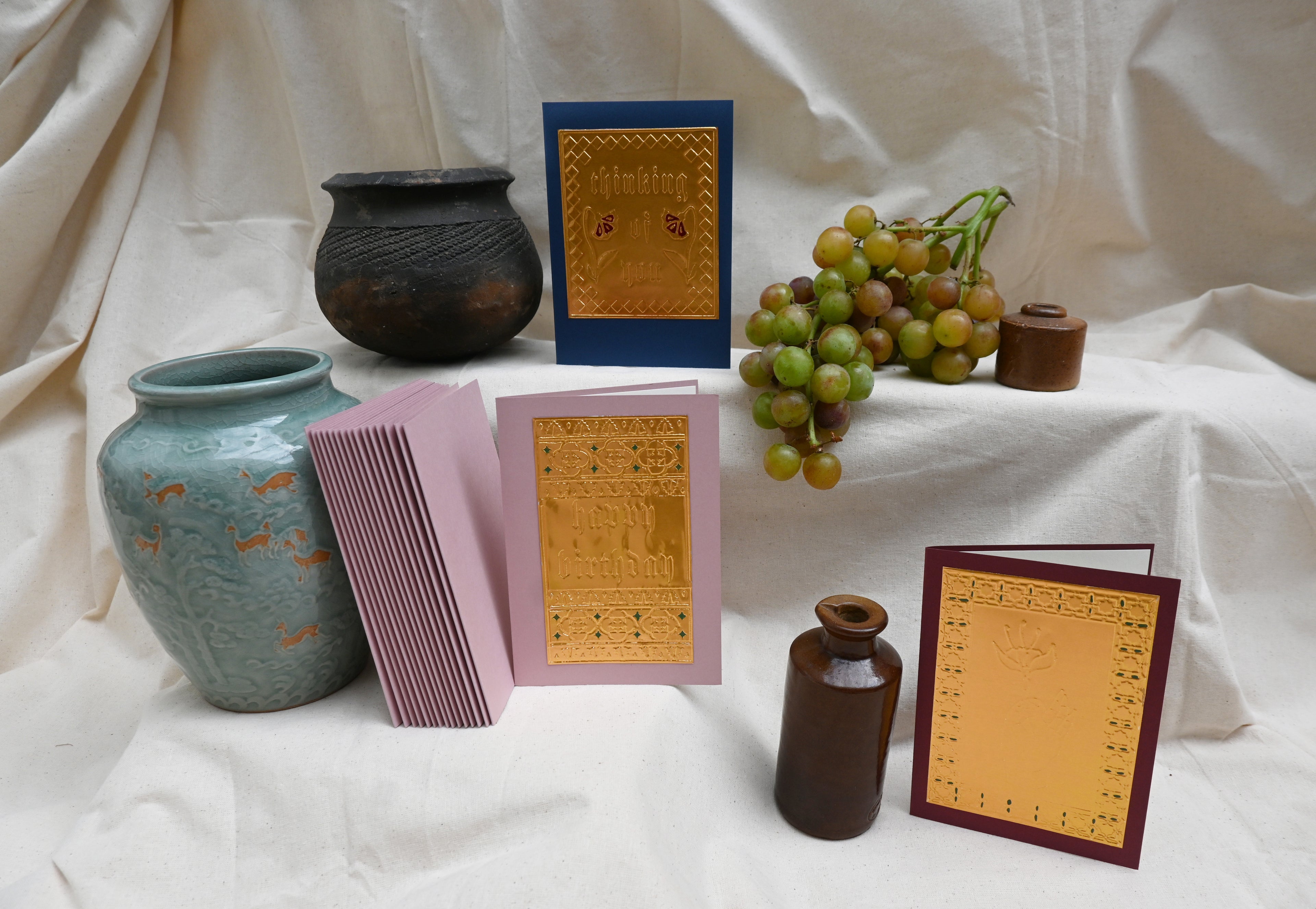 A collection of 3 greeting card by Fabler's Press. Navy blue with the text Thinking Of You. Dusty pink with Happy Birthday and Burgundy card with hand holding a flower. surrounded by ancient looking pots and ceramics and a bunch of green blushing grapes. reminiscent of a Venetian still life on a back drop of cream canvas.