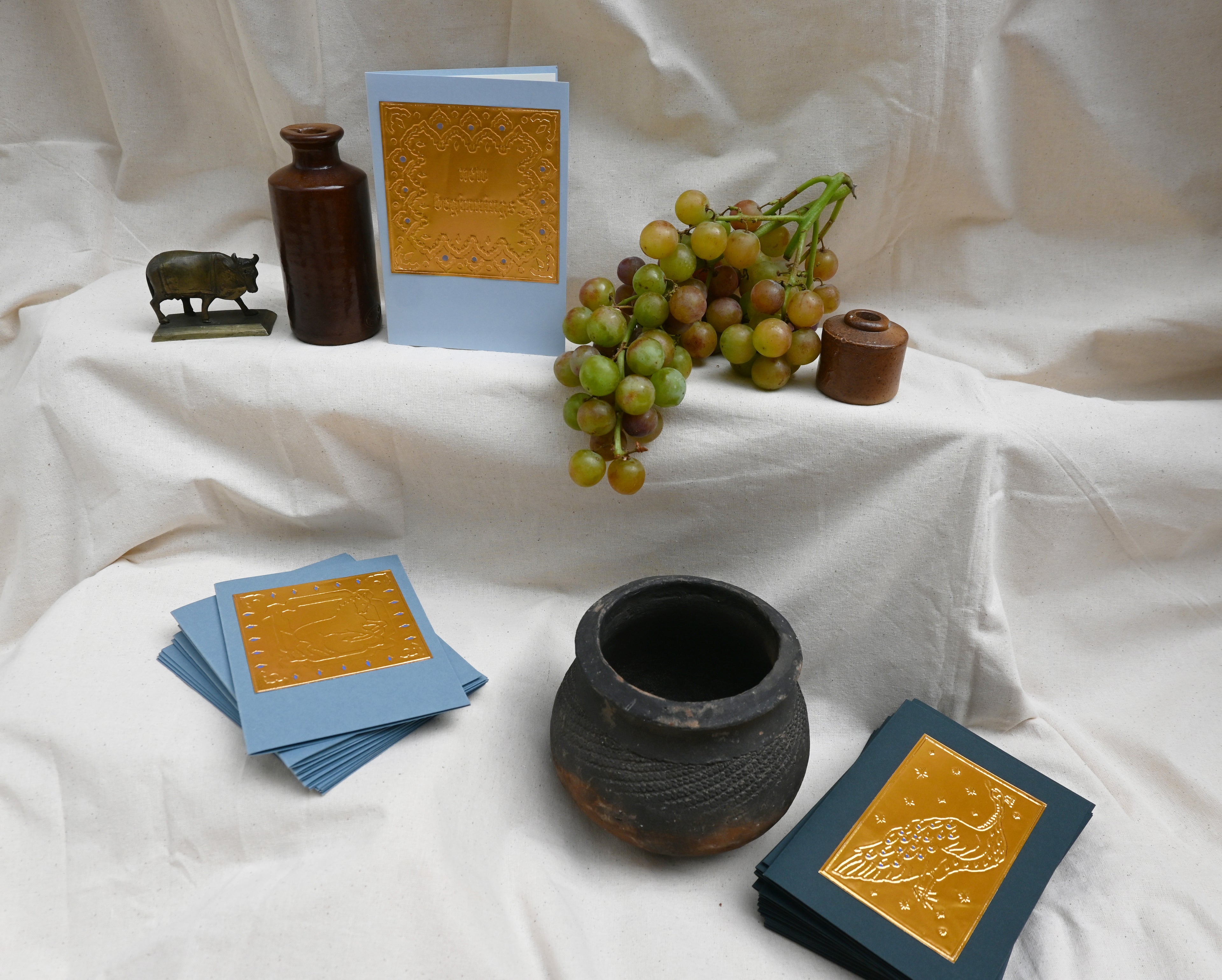 A collection of 3 greeting card by Fabler's Press. Navy blue with peacock Dusty blue with horse and light blue card with text "New Beginnings". surrounded by ancient looking pots bull figurine, and ceramics and a bunch of green blushing grapes. reminiscent of a Venetian still life on a back drop of cream canvas.
