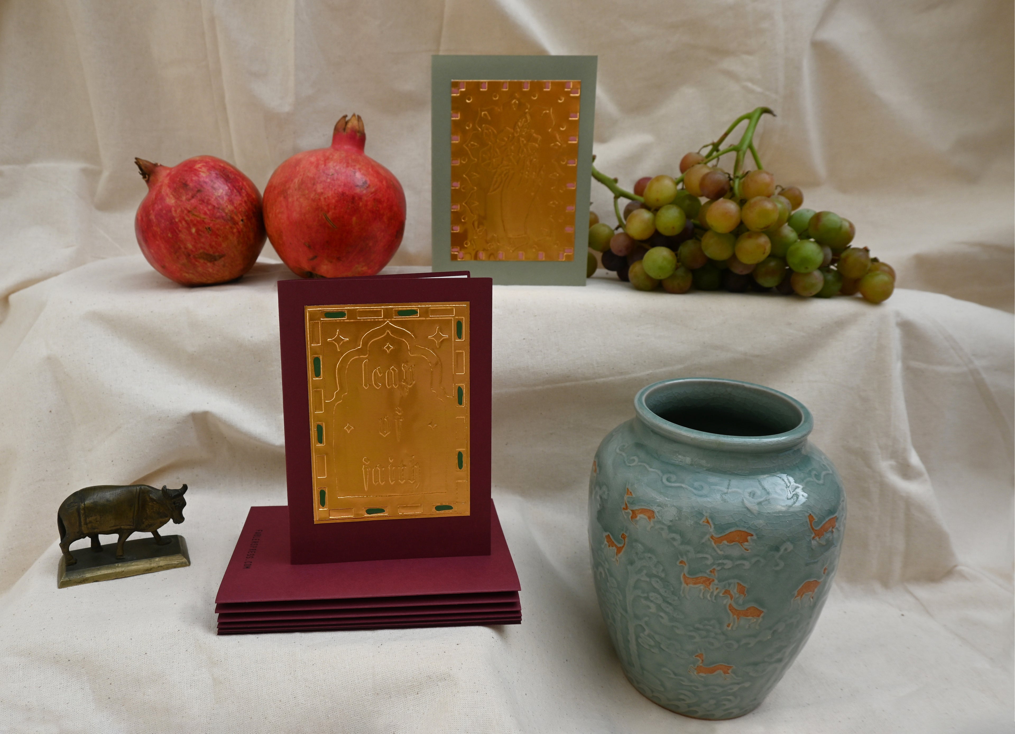 A collection of two greeting card by Fabler's Press. Burgundy with the text Leap Of Faith. and green with a hand holding a bunch of flowers surrounded by ancient looking pots and ceramics, pomegranates, a bull figurine and a bunch of green blushing grapes. reminiscent of a Venetian still life on a back drop of cream canvas.
