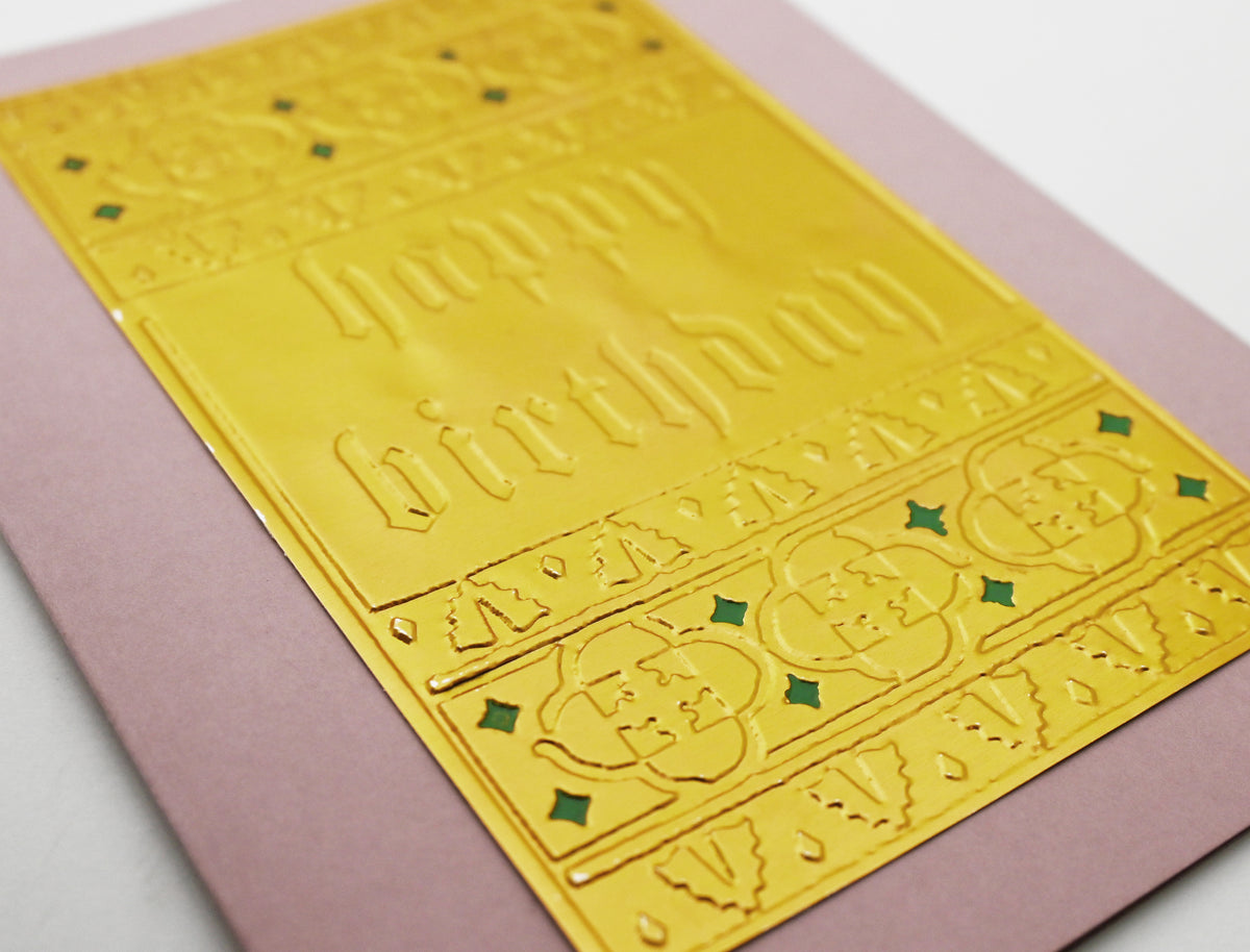 A close-up of a gold foil embossed 'Happy Birthday' card  with ornate border placed on top of a dusty pink  card against a white background.