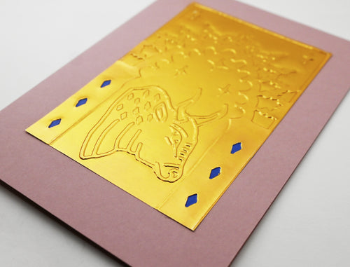 A gold foil card close up featuring an embossed bull design and small blue diamond accents, placed on a dusty pink backing card against a light gray background.