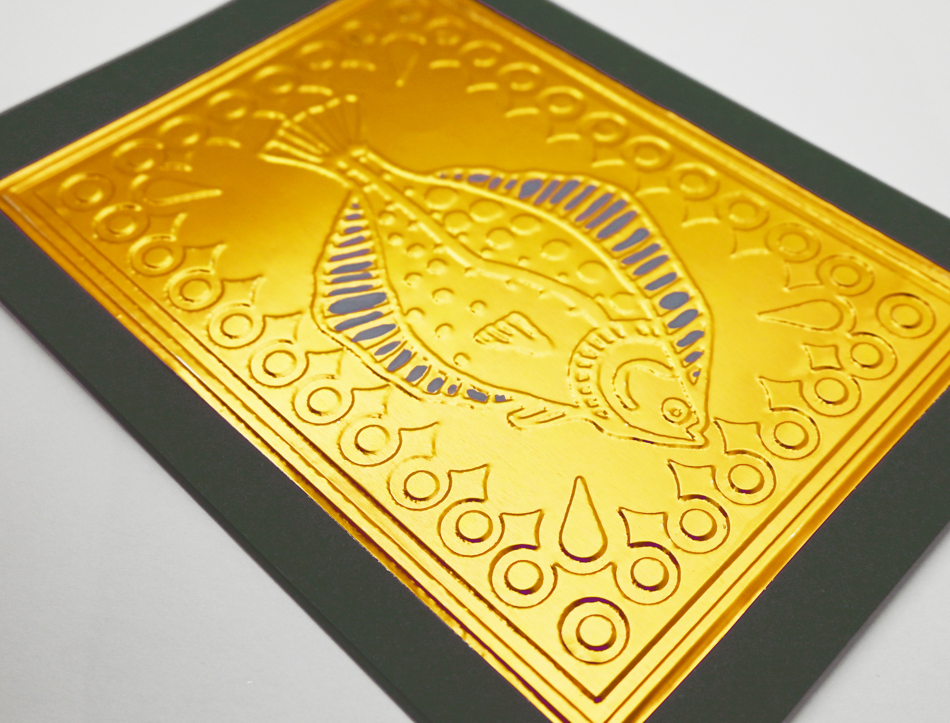 A greeting card close up by fabler's Press with a raised gold fish design on dark green card stock, shown with a plain off-white envelope behind it on a white background.