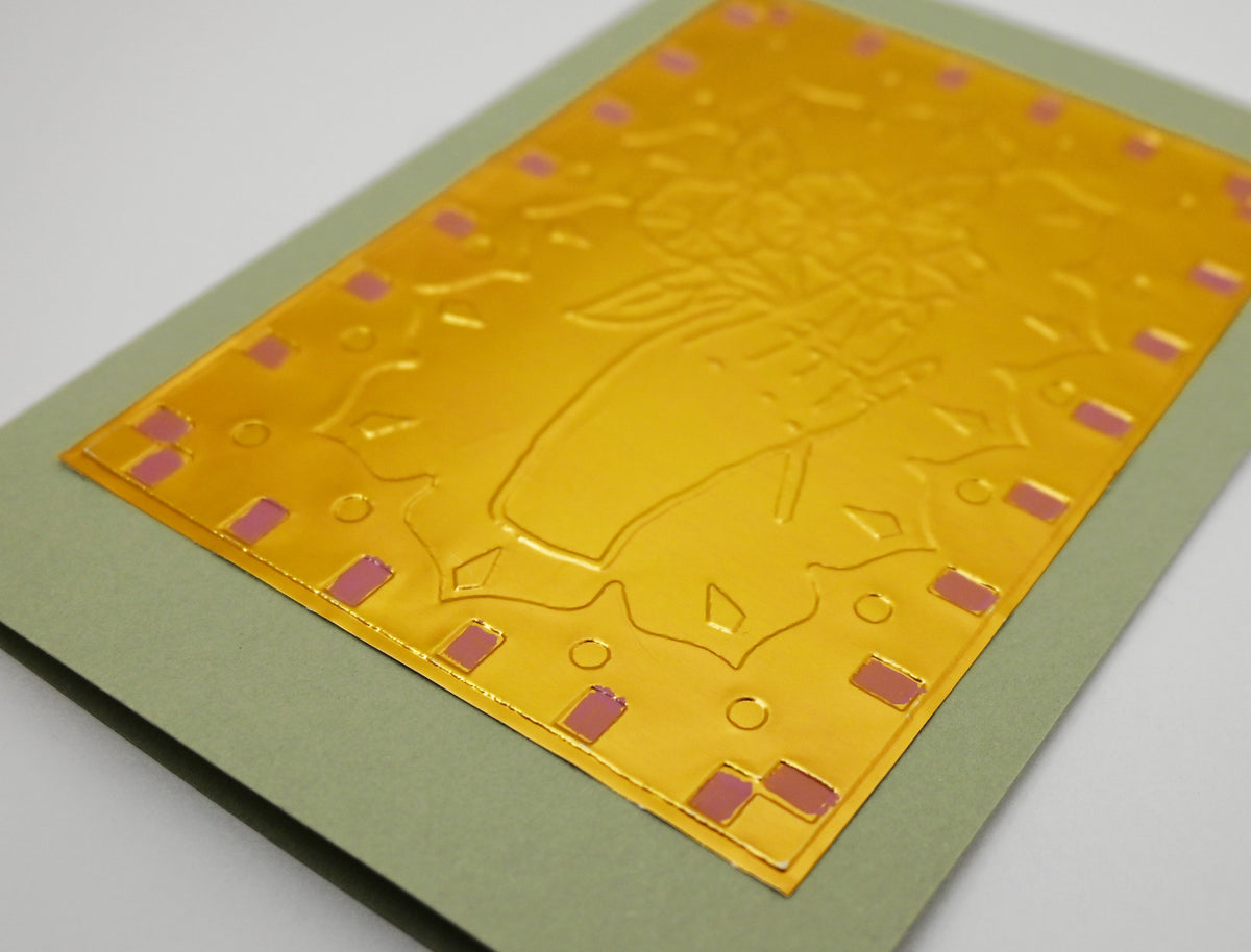 A close-up of a metallic gold decorative card with embossed floral and geometric patterns, mounted on a sage green backing against a light gray background.