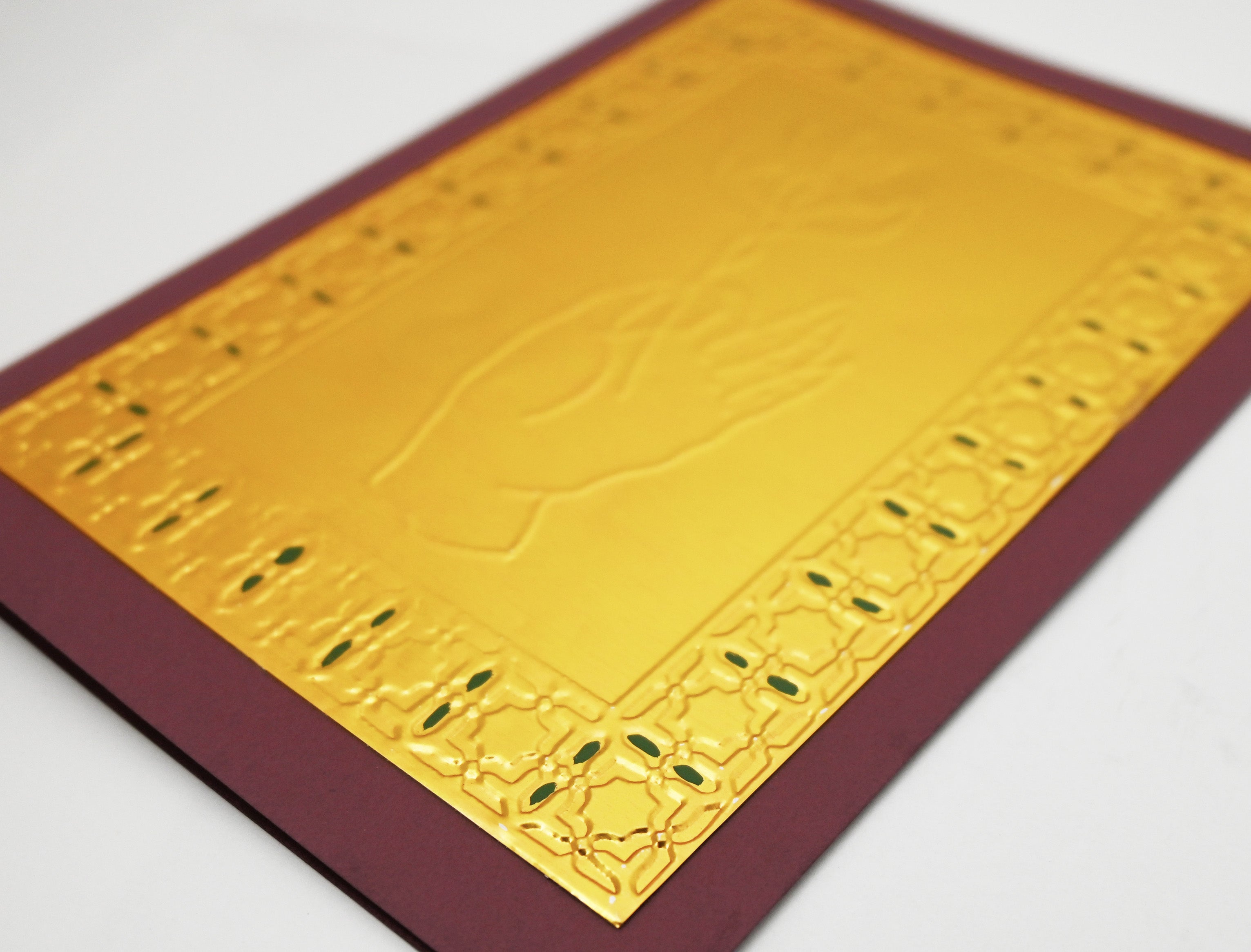 A gold-toned greeting card with a raised hand holding a flower, surrounded by an ornate patterned border, set against a deep burgundy background. Close up shot of the details and the gold pressed foil and painted accents. 