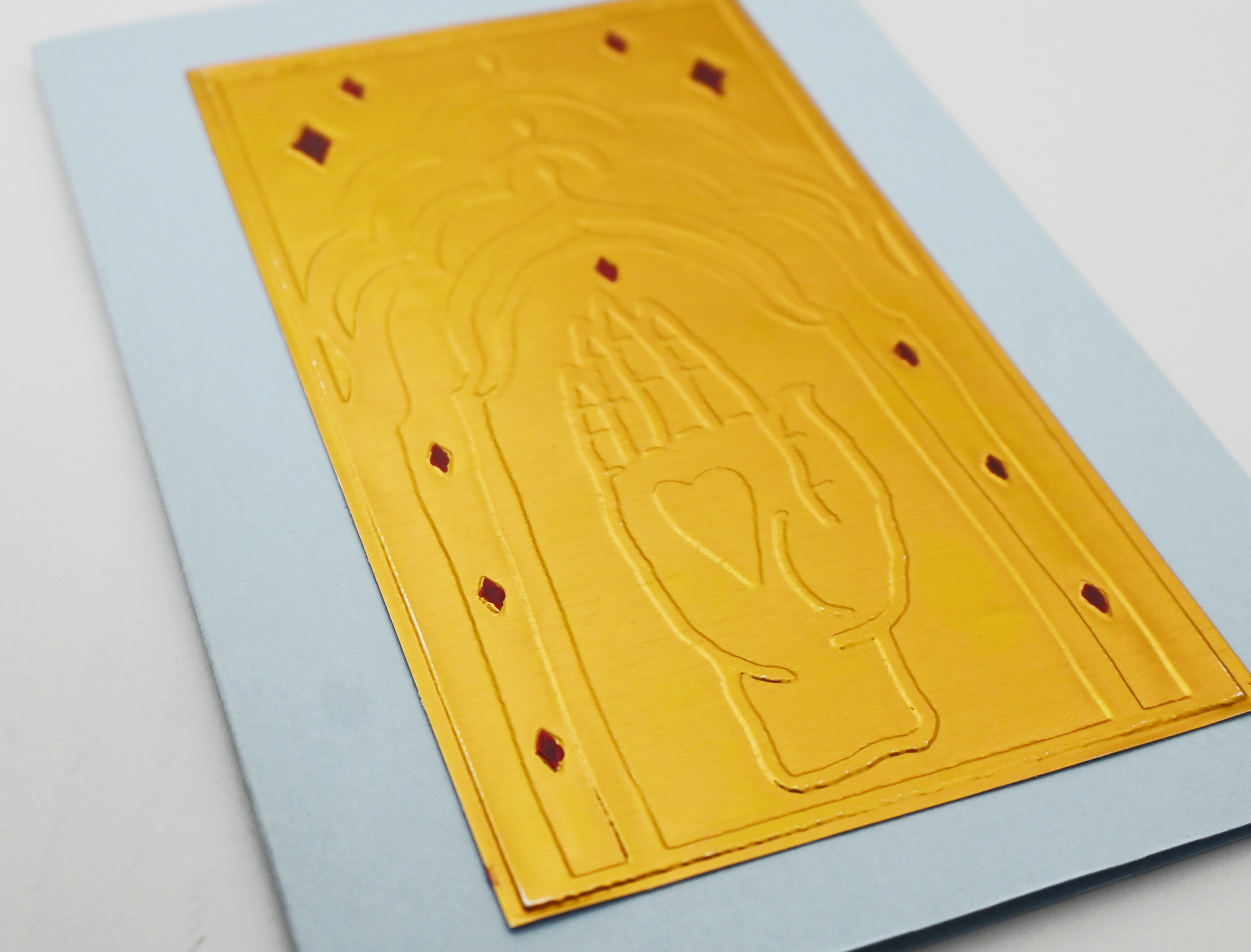 A greeting card close up by fabler's Press with a raised gold hand design on a muted blue cards tock, shown on a white background.