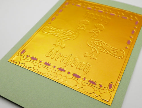 A close-up of a square gold foil card with embossed decorative borders, two birds, and the words 'Happy Birthday,' placed on a light green card against a white background.