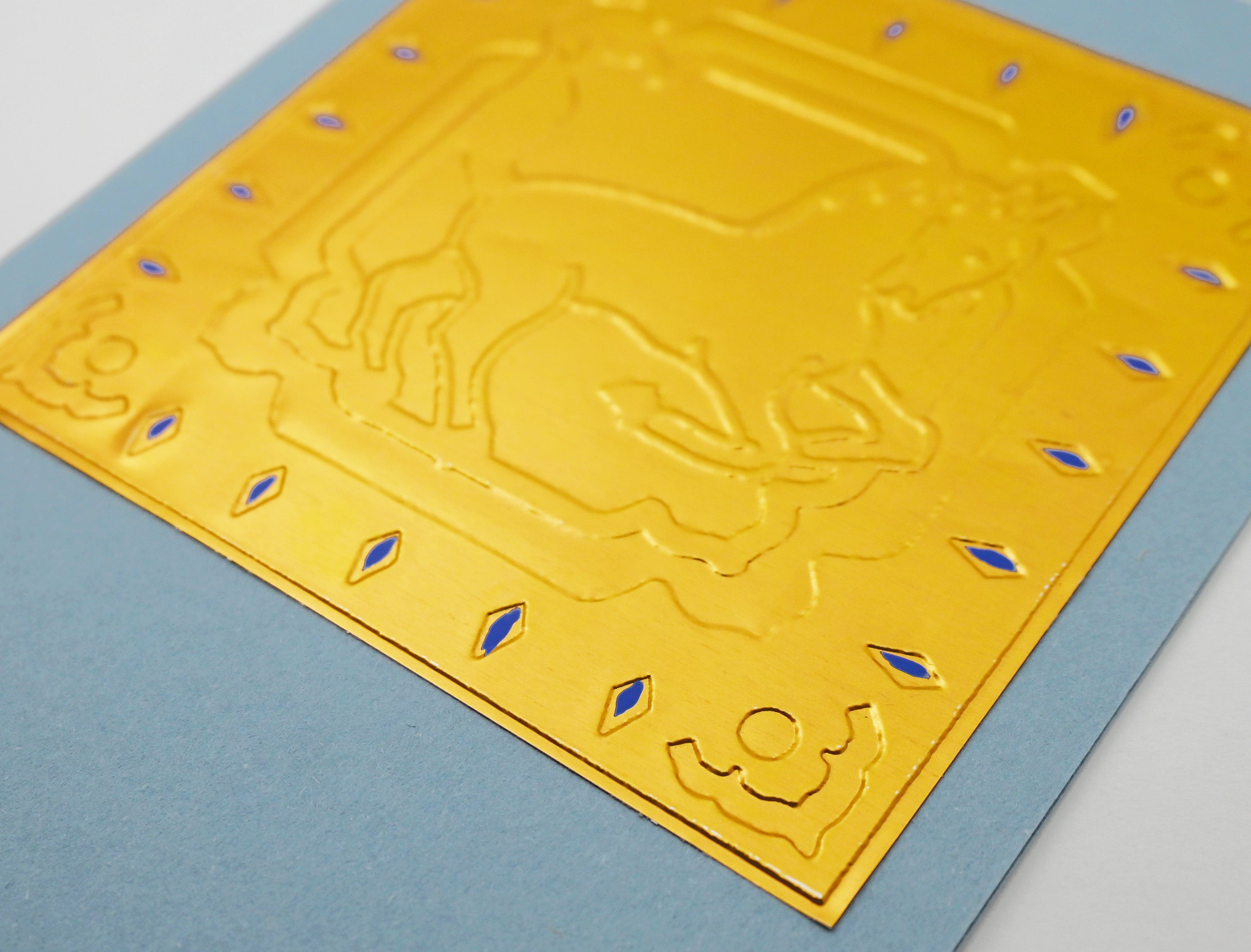 A close-up of a square gold decorative card with embossed horse and diamond motifs, set against a light blue backing card.