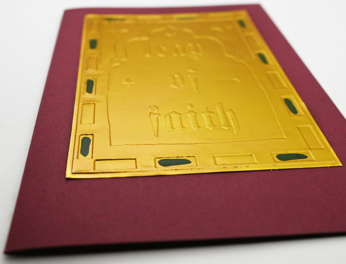 A greeting card close up with a deep burgundy border and a central gold foil panel embossed with the words 'leap of faith,' shown with a plain cream envelope behind it on a white background.