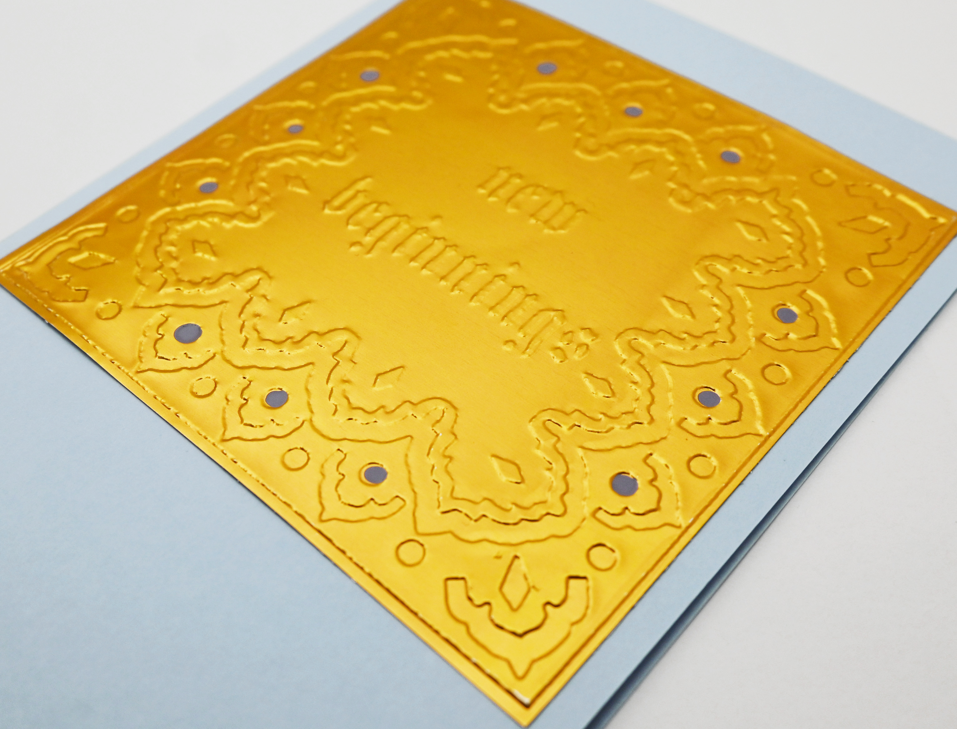 A greeting card close up by fabler's Press with a raised text design "New Beginnings" with and ornate border on a muted blue card stock, shown with a plain off-white envelope behind it on a white background.