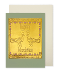 A greeting card with a metallic gold front featuring two peacock illustrations and the words 'happy birthday' in gothic lettering, set against a muted sage green border. A cream-colored envelope is partially visible behind it on a white background.