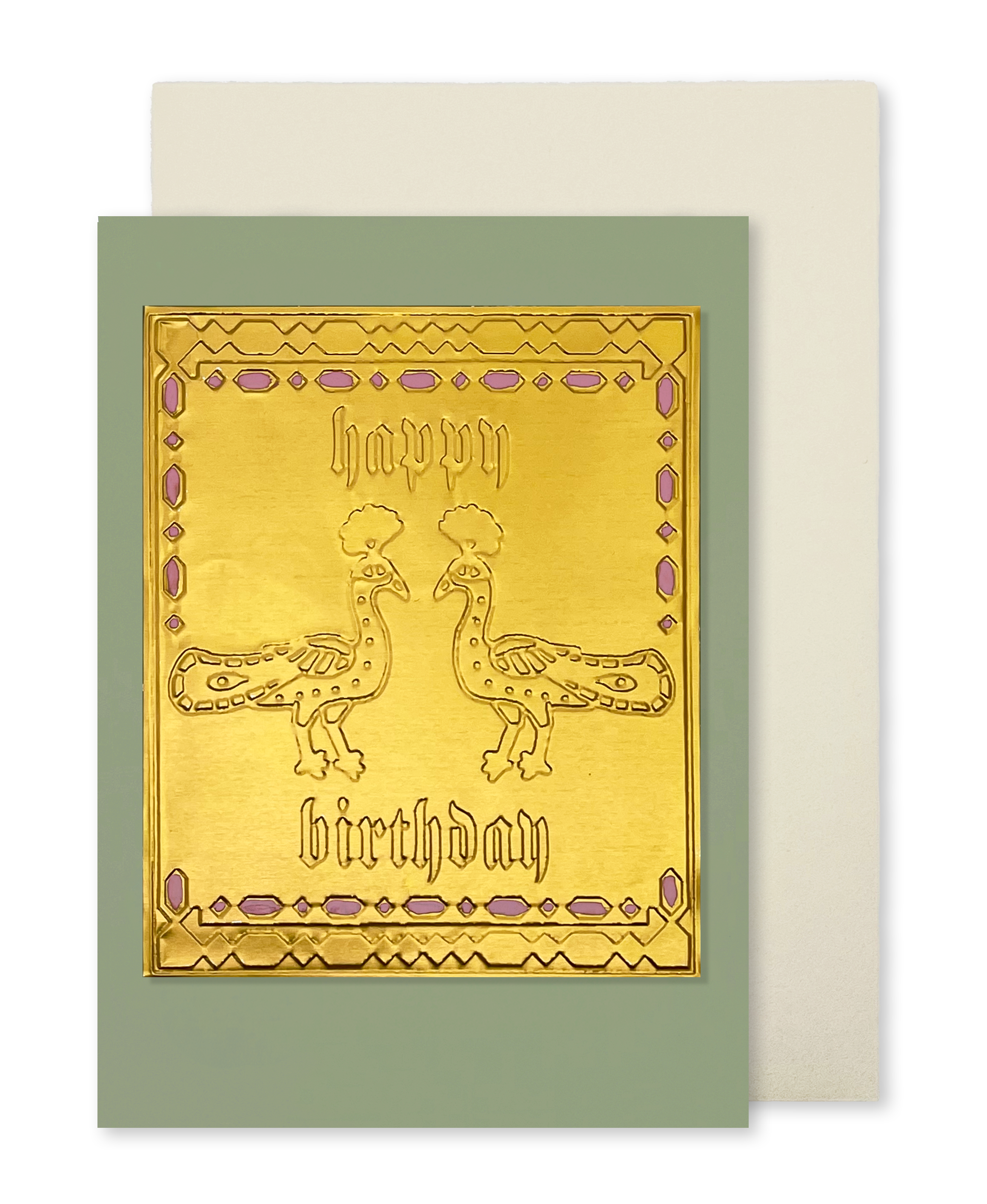 A greeting card with a metallic gold front featuring two peacock illustrations and the words 'happy birthday' in gothic lettering, set against a muted sage green border. A cream-colored envelope is partially visible behind it on a white background.