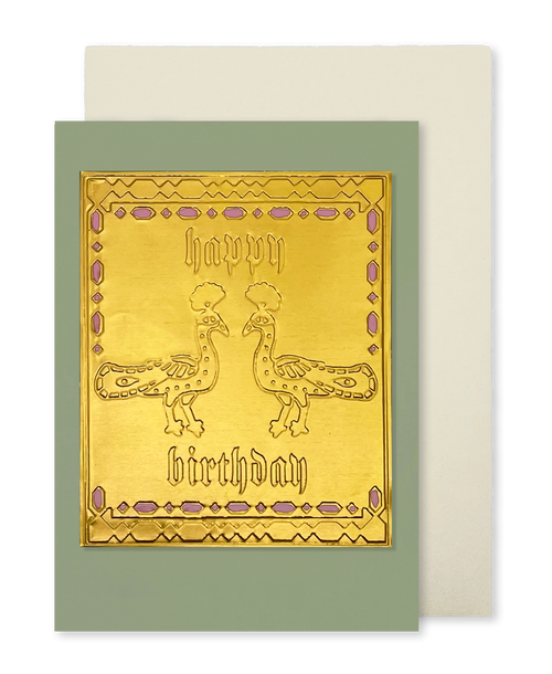 A greeting card with a metallic gold front featuring two peacock illustrations and the words 'happy birthday' in gothic lettering, set against a muted sage green border. A cream-colored envelope is partially visible behind it on a white background.