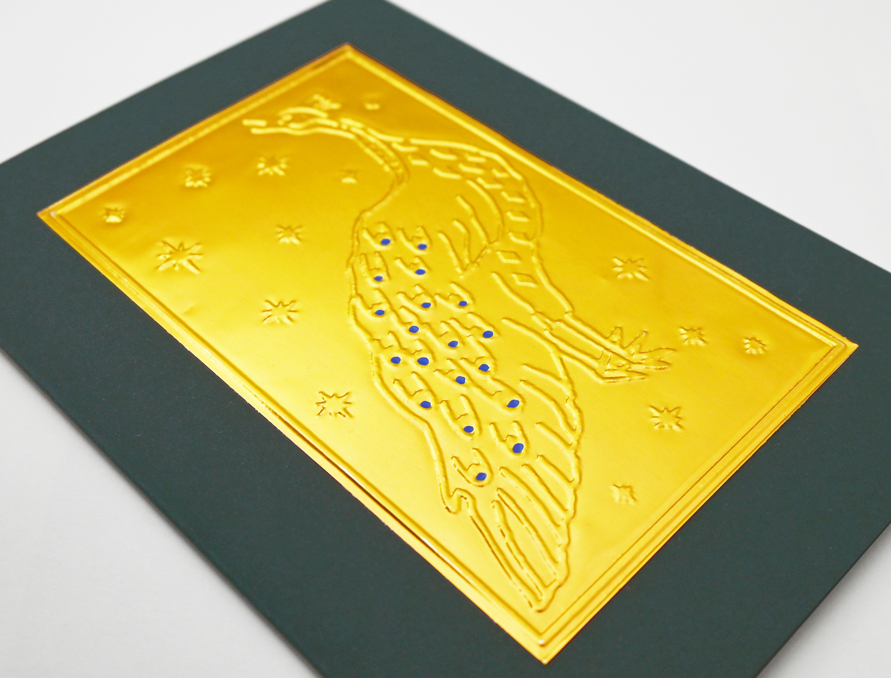 A greeting card close up with a dark green border and a central gold foil embossed peacock design with blue jewel accents on an off-white background.