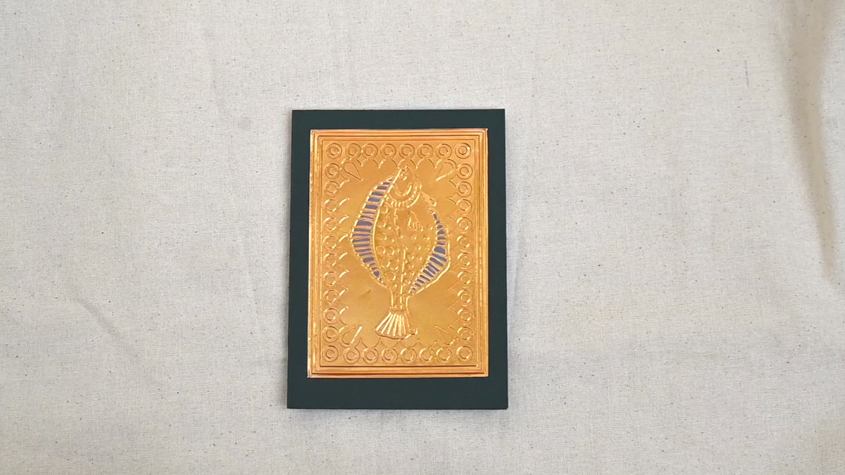 A video of a gold-toned embossed greeting card with a fish surrounded by an ornate patterned border, set against a deep dark green card. Two hands picking it up, opening it to show the ivory white insert, closing it and putting it down. All on a canvas background.