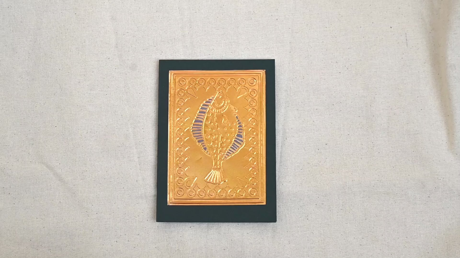 A video of a gold-toned embossed greeting card with a fish surrounded by an ornate patterned border, set against a deep dark green card. Two hands picking it up, opening it to show the ivory white insert, closing it and putting it down. All on a canvas background.