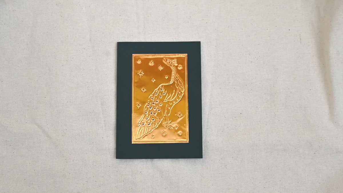 A video of a gold-toned embossed greeting card with a peacock surrounded by an star patterned background and jewel tone blue paint, set against a deep green card. Two hands picking it up, opening it to show the ivory white insert, closing it and putting it down. All on a canvas background.
