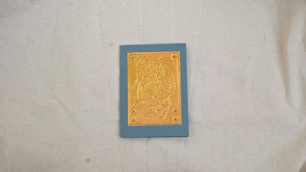 A video of a gold-toned embossed greeting card with a tiger motif, set against a dusty blue card. Two hands picking it up, opening it to show the ivory white insert, closing it and putting it down. All on a canvas background.

