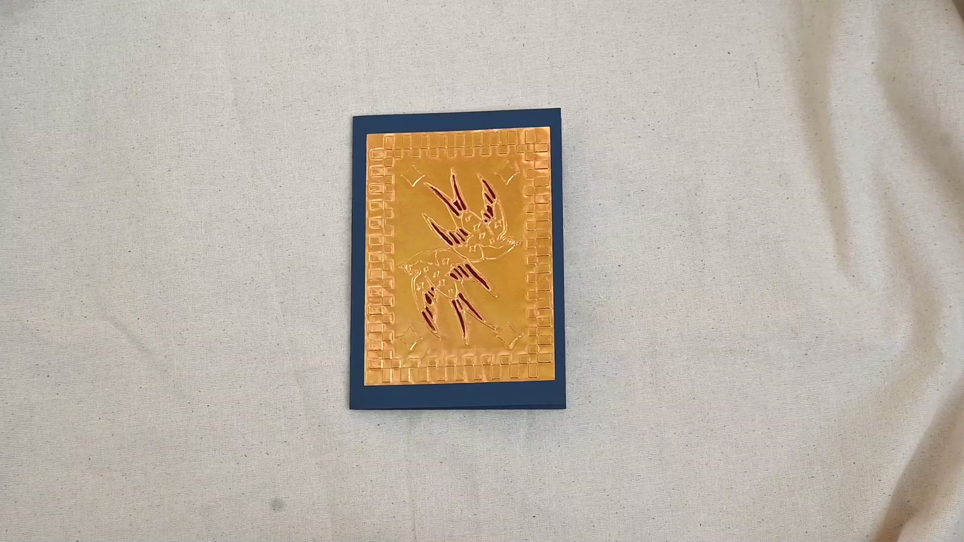 A video of a gold-toned embossed greeting card with two swallows, surrounded by a square patterned border, set against a navy blue card. Two hands picking it up, opening it to show the ivory white insert, closing it and putting it down. All on a canvas background.

