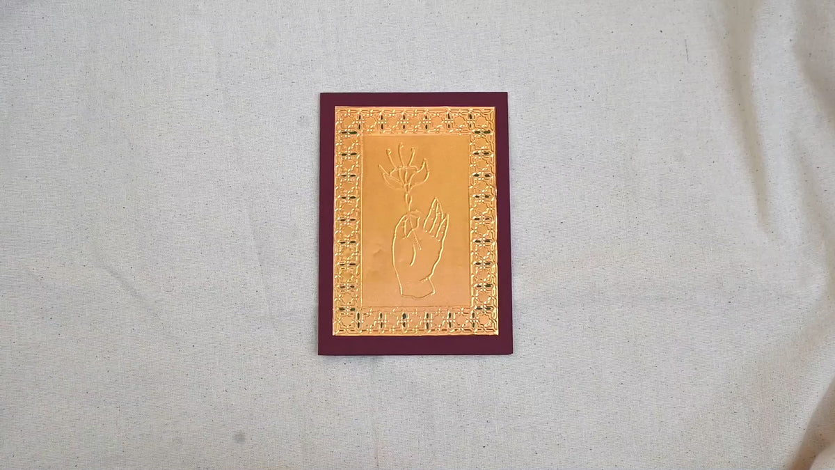 A video of a gold-toned greeting card with a raised hand holding a flower, surrounded by an ornate patterned border, set against a deep burgundy card. Two hands picking it up, opening it to show the ivory white insert, closing it and putting it down. All on a canvas background.
