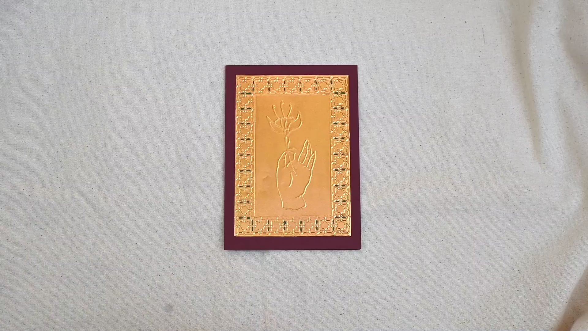 A video of a gold-toned greeting card with a raised hand holding a flower, surrounded by an ornate patterned border, set against a deep burgundy card. Two hands picking it up, opening it to show the ivory white insert, closing it and putting it down. All on a canvas background.