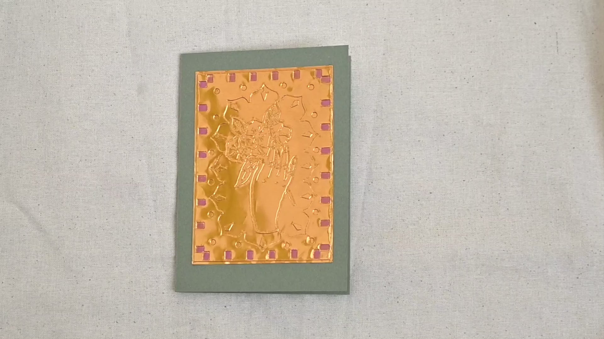 A video of a gold-toned embossed greeting card with a hand holding a flowers, surrounded by an ornate patterned border, set against a sage green card. Two hands picking it up, opening it to show the ivory white insert, closing it and putting it down. All on a canvas background.