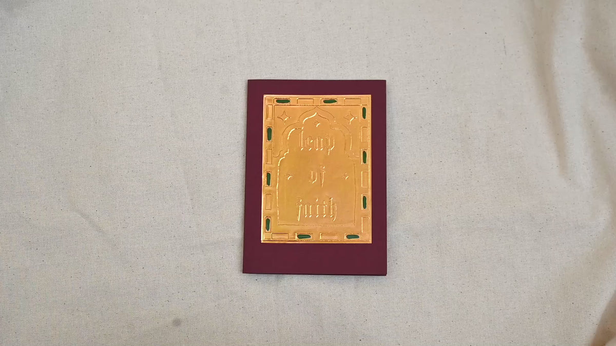A video of a gold-toned embossed greeting card with text "Leap Of Faith", surrounded by an ornate patterned border, set against a deep burgundy card. Two hands picking it up, opening it to show the ivory white insert, closing it and putting it down. All on a canvas background.

