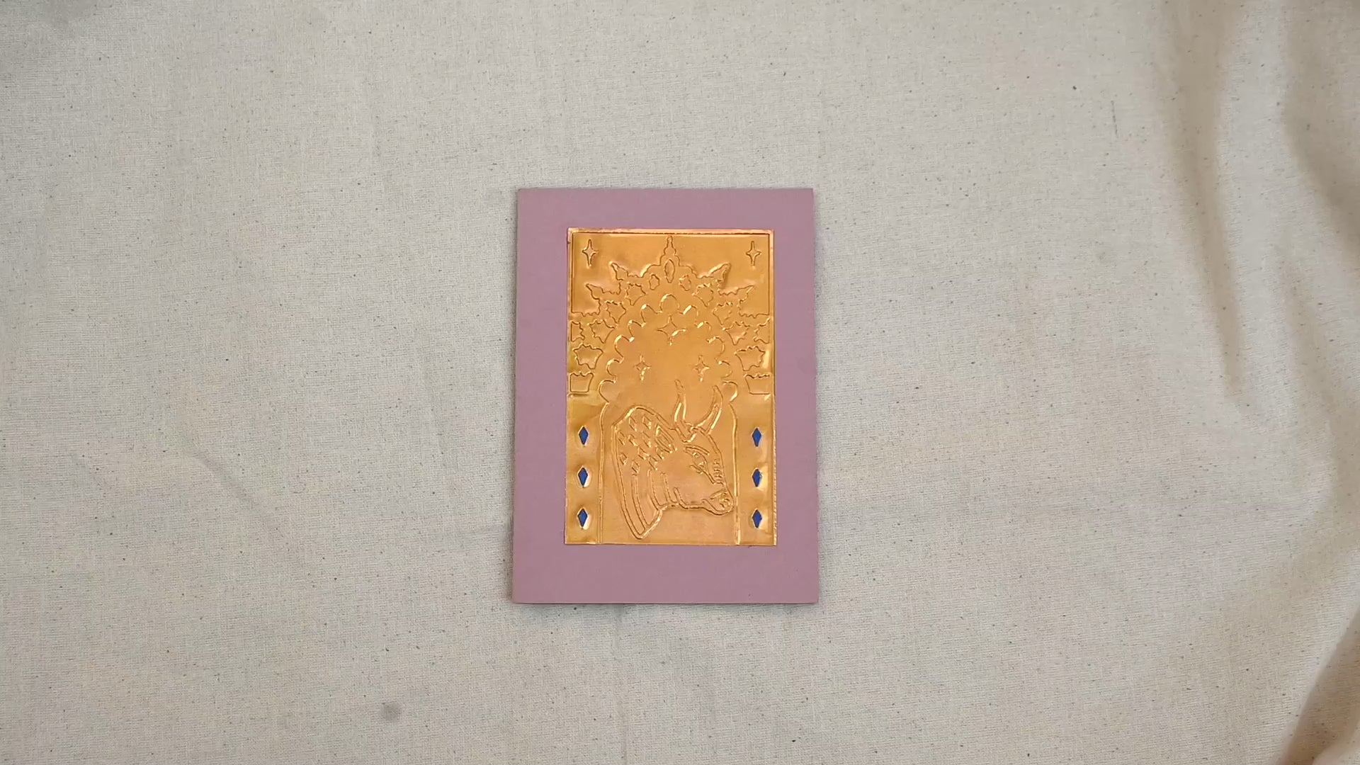 A video of a gold-toned embossed greeting card with a raised bull, surrounded by an ornate patterned border, set against a dusty pink card. Two hands picking it up, opening it to show the ivory white insert, closing it and putting it down. All on a canvas background.
