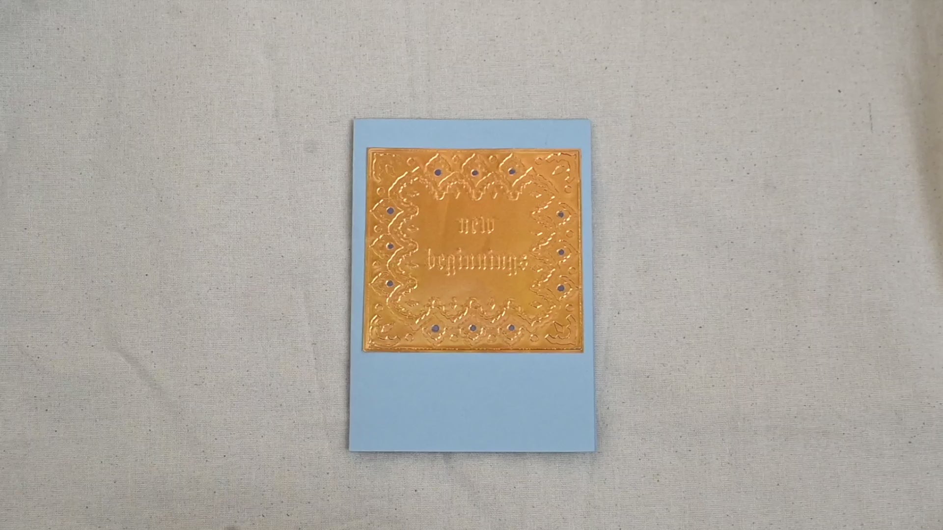 A video of a gold-toned embossed greeting card with text "New Beginnings" surrounded by an ornate patterned border, set against a light blue card. Two hands picking it up, opening it to show the ivory white insert, closing it and putting it down. All on a canvas background.
