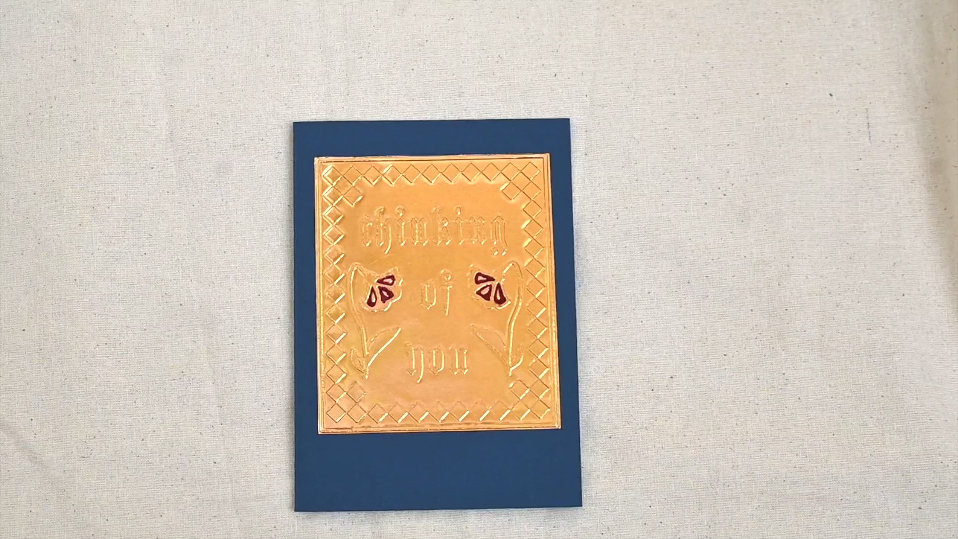 A video of a gold-toned embossed greeting card with text "Thinking Of You" surrounded by 2 red flowers and an square patterned border, set against a deep navy blue card. Two hands picking it up, opening it to show the ivory white insert, closing it and putting it down. All on a canvas background.