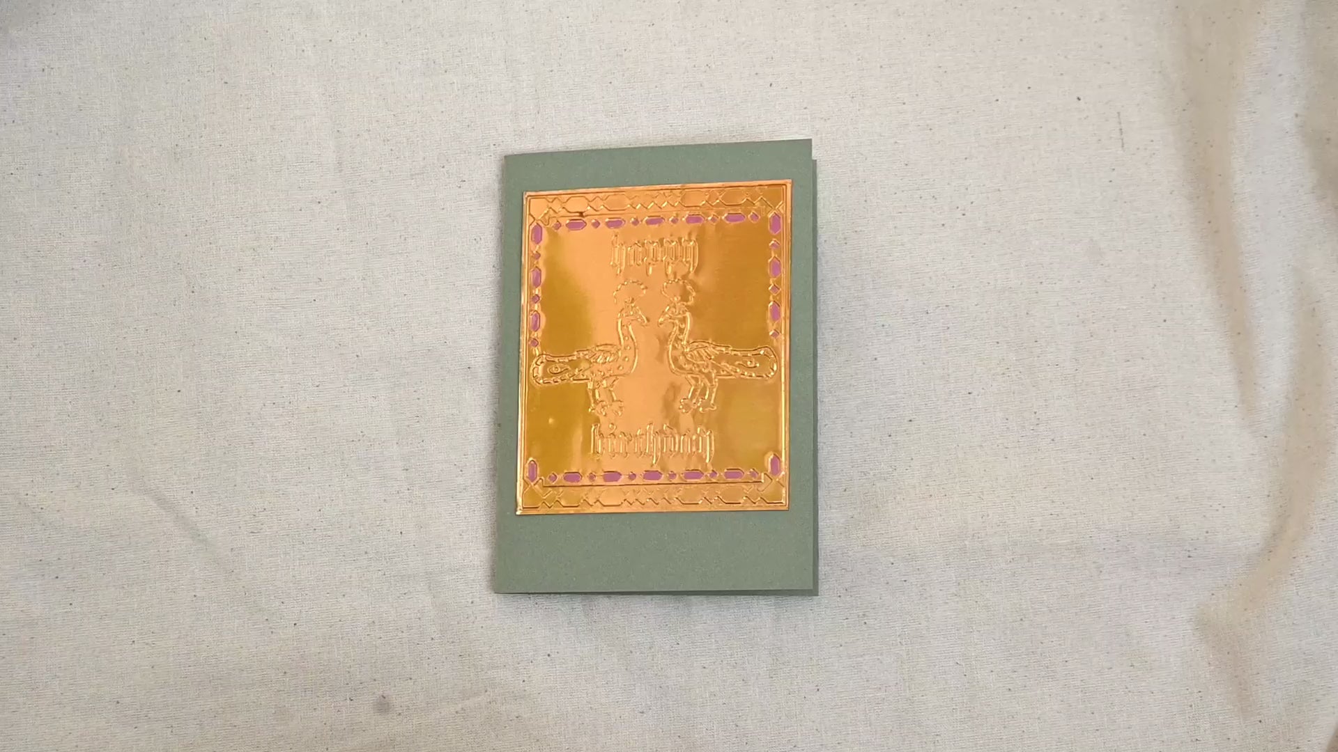 A video of a gold-toned embossed greeting card with two peococks and Happy Birthday text, surrounded by an ornate patterned border, set against a sage green card. Two hands picking it up, opening it to show the ivory white insert, closing it and putting it down. All on a canvas background.