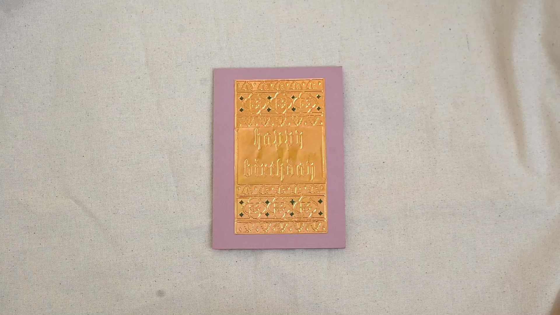 A video of a gold-toned embossed greeting card with text "Happy Birthday" surrounded by an ornate patterned border, set against a dusty pink card. Two hands picking it up, opening it to show the ivory white insert, closing it and putting it down. All on a canvas background.