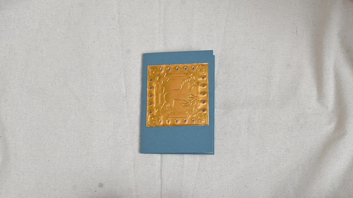 A video of a gold-toned embossed greeting card with a horse surrounded by an ornate patterned border, set against a blue card. Two hands picking it up, opening it to show the ivory white insert, closing it and putting it down. All on a canvas background.
