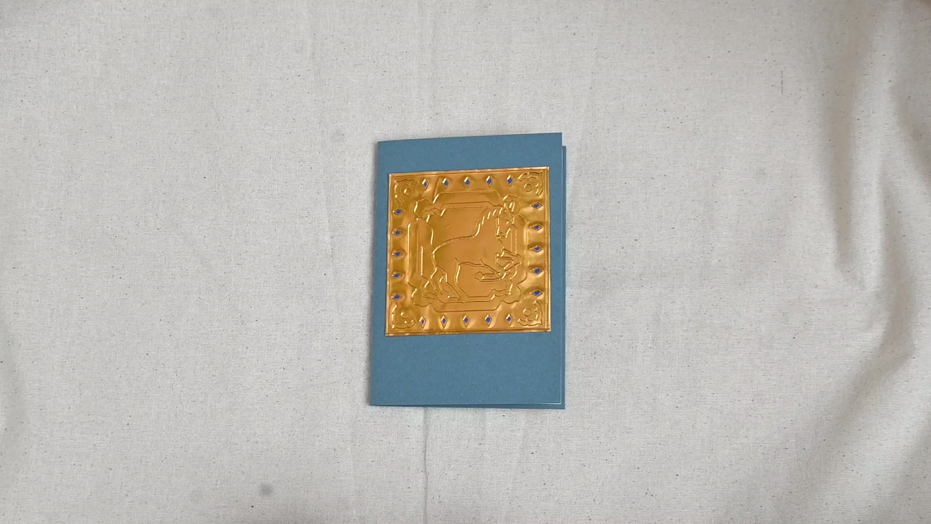 A video of a gold-toned embossed greeting card with a horse surrounded by an ornate patterned border, set against a blue card. Two hands picking it up, opening it to show the ivory white insert, closing it and putting it down. All on a canvas background.
