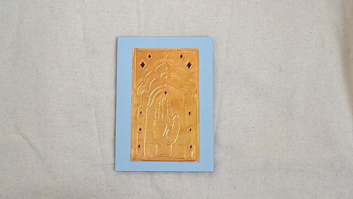 A video of a gold-toned embossed greeting card with a raised hand holding a heart, surrounded by an ornate patterned border, set against a blue card. Two hands picking it up, opening it to show the ivory white insert, closing it and putting it down. All on a canvas background.
