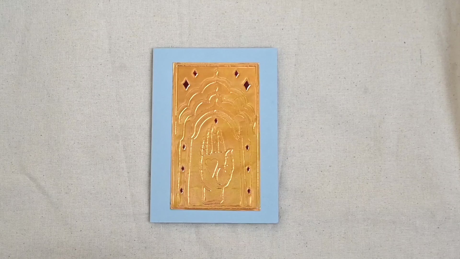 A video of a gold-toned embossed greeting card with a raised hand holding a heart, surrounded by an ornate patterned border, set against a blue card. Two hands picking it up, opening it to show the ivory white insert, closing it and putting it down. All on a canvas background.
