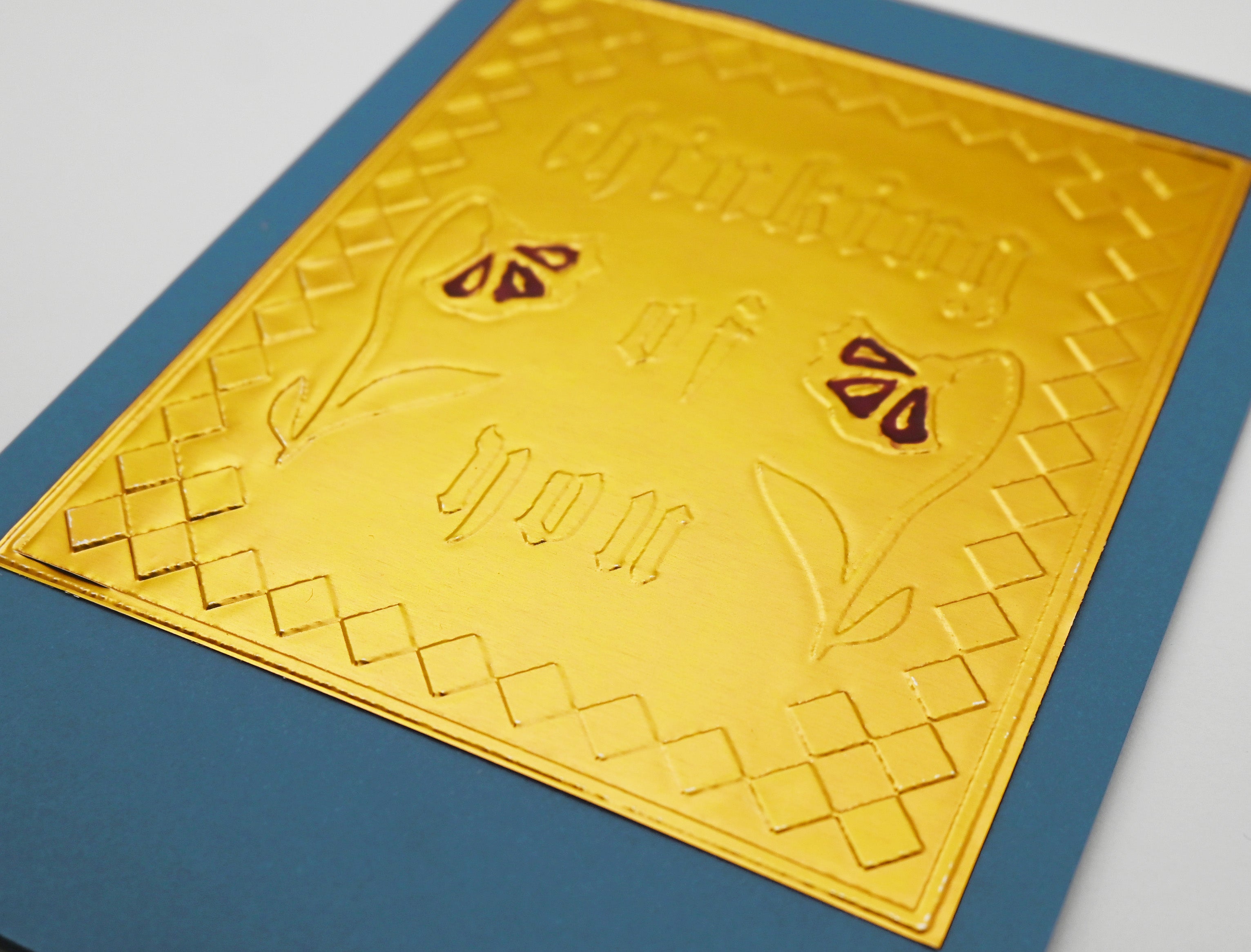 A close-up of a square gold foil card with embossed decorative text, Thinking Of You, and floral motifs, layered over a blue card on a off white background.