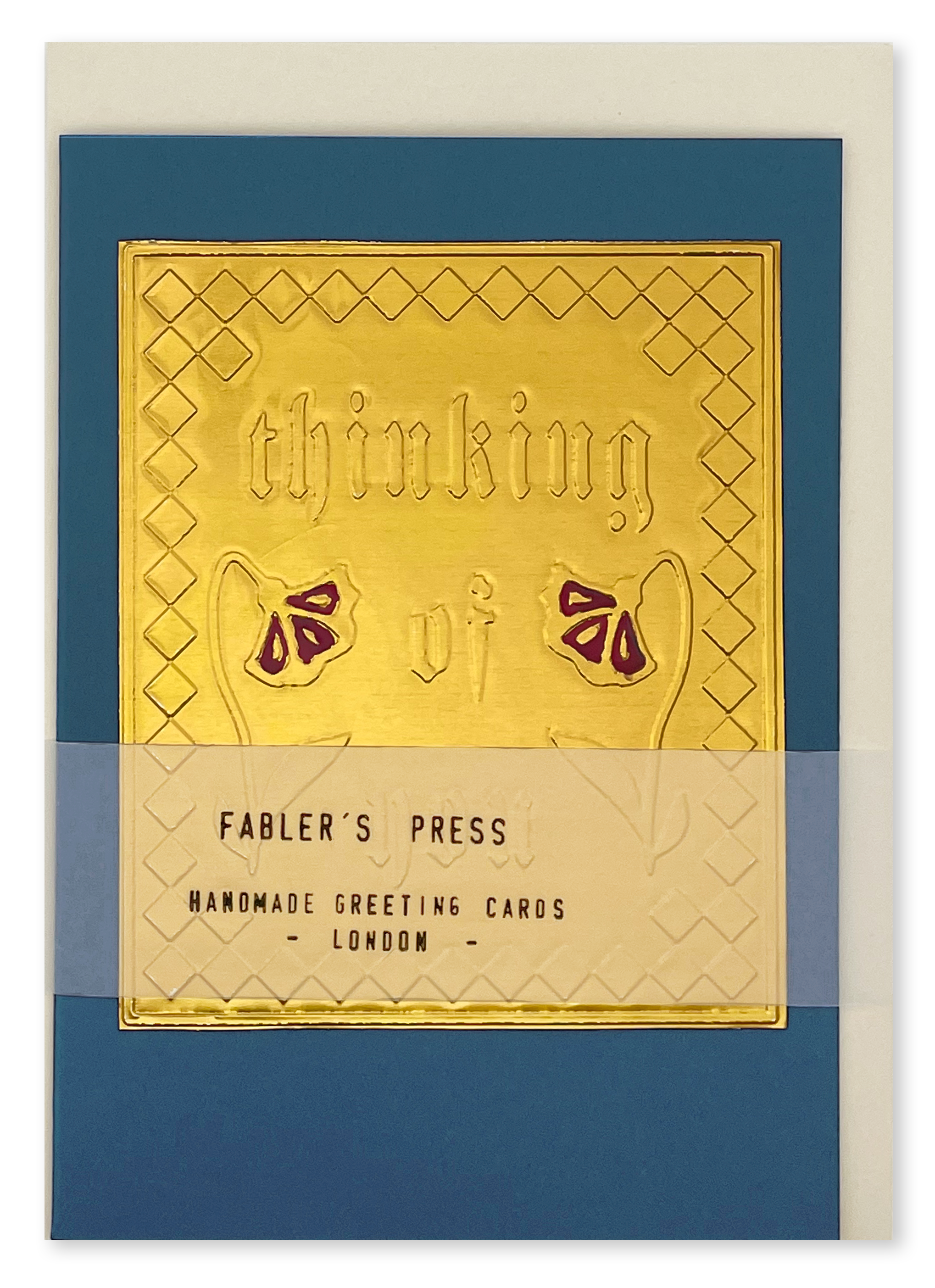 A Fabler's Press handmade greeting card with a gold foil embossed design reading 'thinking of You' and flower motifs on a blue background, shown with a cream envelope behind it. wrapped with a translucent band labeled 'FABLER'S PRESS HANDMADE GREETING CARDS LONDON'.