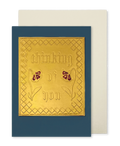 A navy blue greeting card with a raised gold foil panel reading 'thinking of you' and small red flower motifs, shown with a cream envelope 