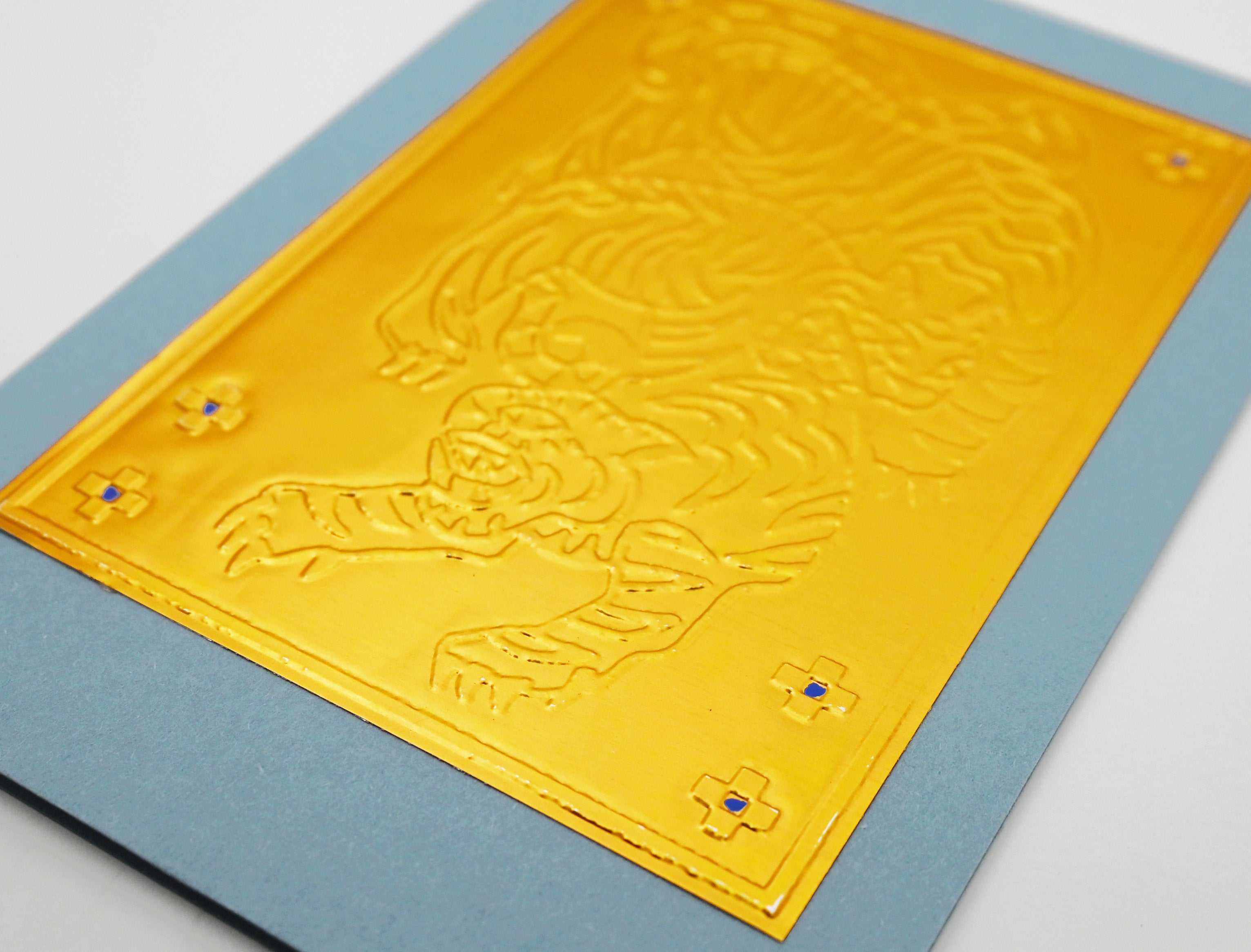 A close-up of gold embossed metal featuring a tiger design, mounted on a light blue card against a white background.
